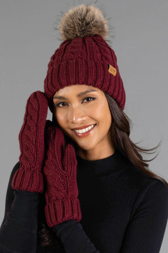 A woman smiles while wearing the Panache Apparel Collection Burgundy Cable Knit Design Pom Hat with a faux fur pom-pom, paired with matching knit mittens and a black sweater, posing against a plain gray background.