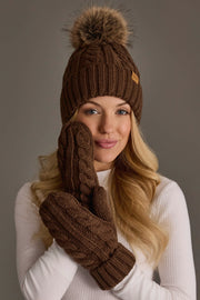 A woman in a white sweater wears the Panache Apparel Collection’s Brown Cable Knit Design Pom Hat, along with matching brown mittens, holding one mittened hand up to her face as she smiles softly at the camera.