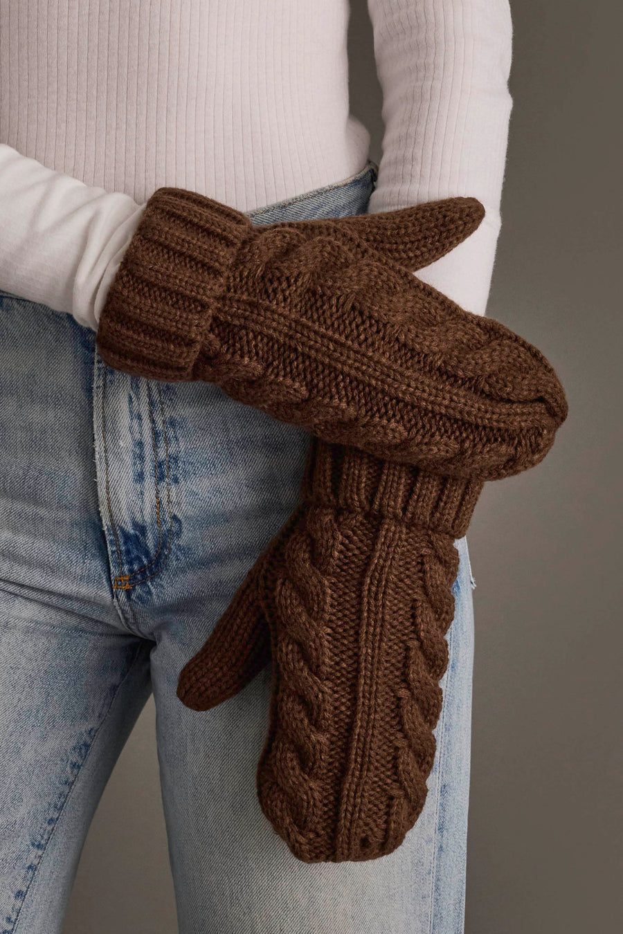 A person dressed in light blue jeans and a white ribbed sweater holds the Brown Cable Knit Design Mittens by Panache Apparel Collection.