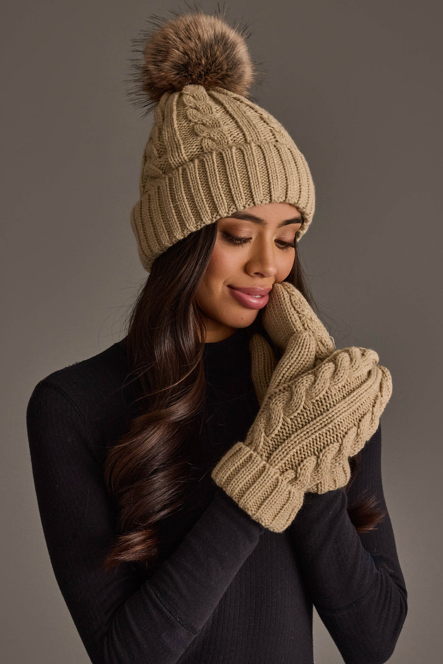 A woman wearing the Panache Apparel Collection Tan Cable Knit Design Pom Hat and matching knit mittens, paired with a black top, poses with a gentle smile against a plain background.