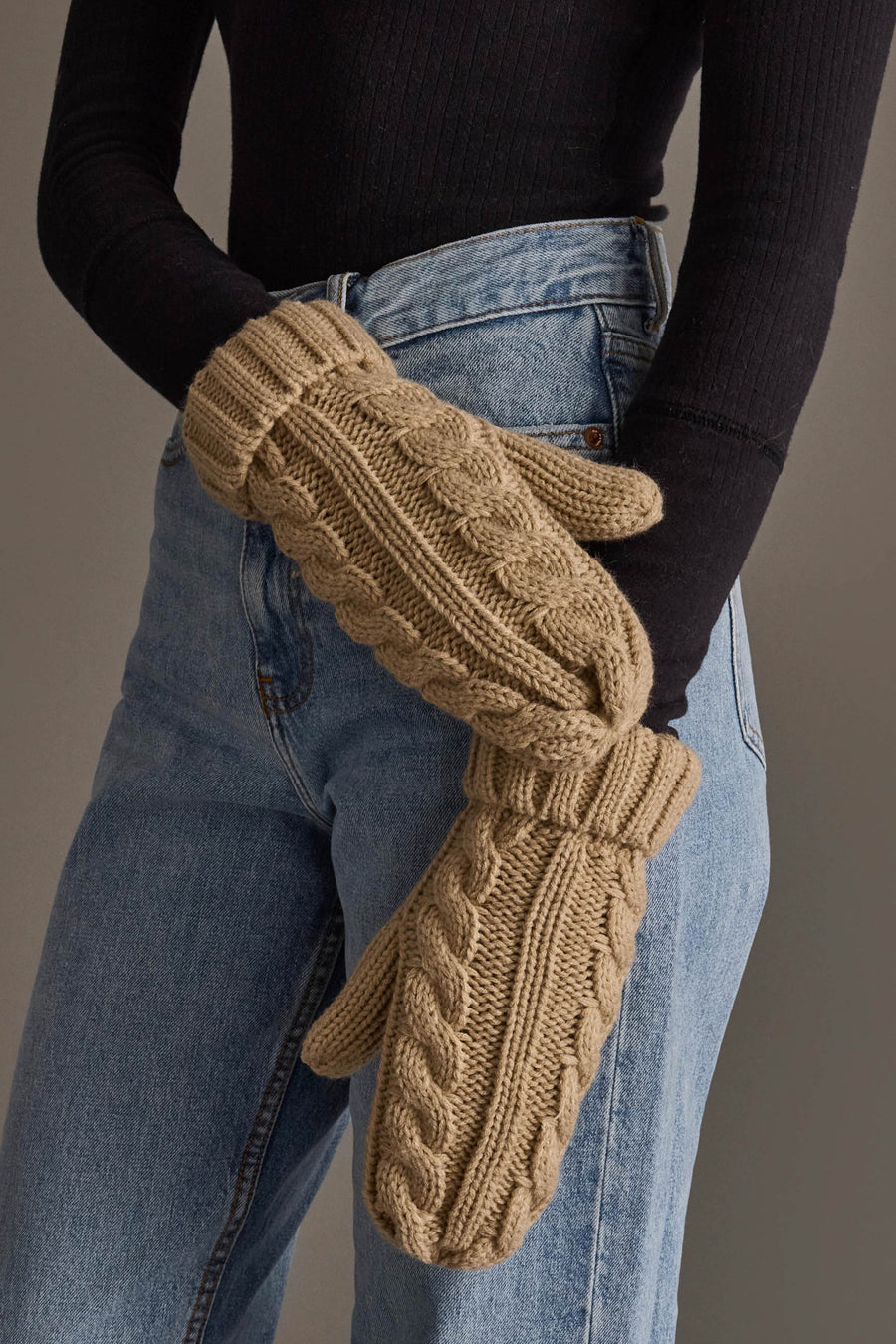 A person wearing a black ribbed long-sleeve top, blue jeans, and the Panache Apparel Collection Tan Cable Knit Design Mittens stands with arms crossed.