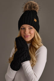 A woman with long blonde hair wears the Black Cable Knit Design Pom Hat from Panache Apparel Collection, featuring a fur pom-pom and matching black mittens. She is dressed in a white ribbed long-sleeve top and smiles softly against a plain gray background.