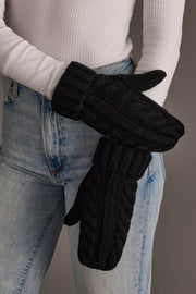 A person wearing blue jeans and a white ribbed sweater holds their arms crossed, showing Black Cable Knit Design Mittens by Panache Apparel Collection.