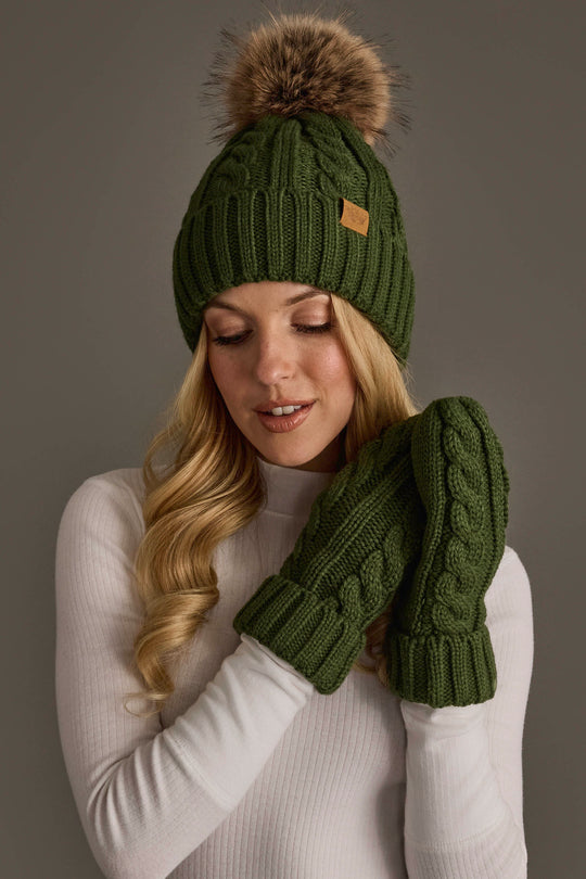 A woman wearing the Panache Apparel Collection’s Dark Green Cable Knit Design Pom Hat, paired with matching green mittens and a white sweater, poses against a neutral background.
