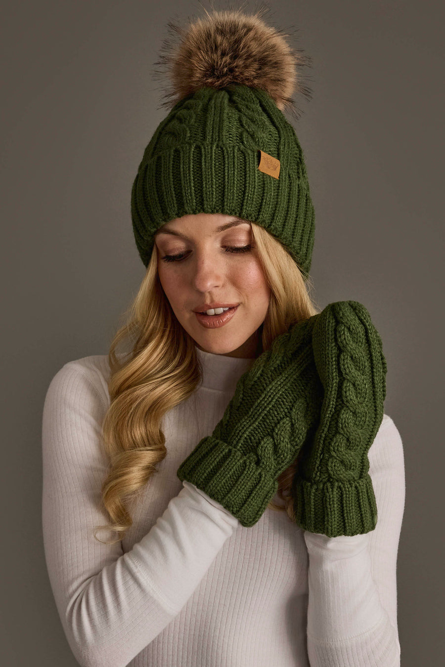 A woman wearing the Panache Apparel Collection’s Dark Green Cable Knit Design Pom Hat, paired with matching green mittens and a white sweater, poses against a neutral background.