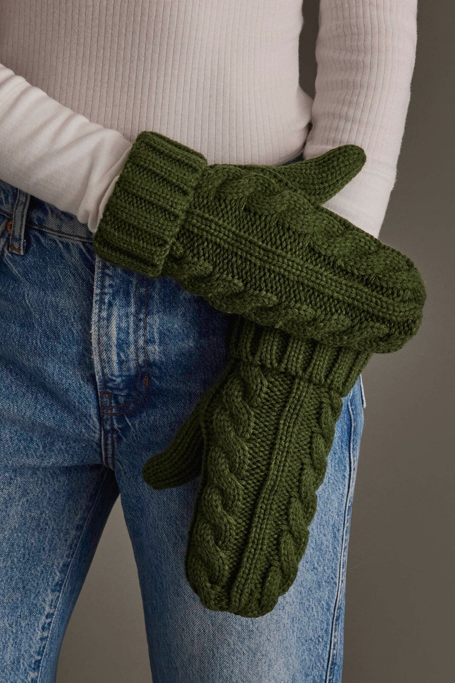 A person wearing a white ribbed sweater and blue jeans holds a pair of Panache Apparel Collection Dark Green Cable Knit Design Mittens.