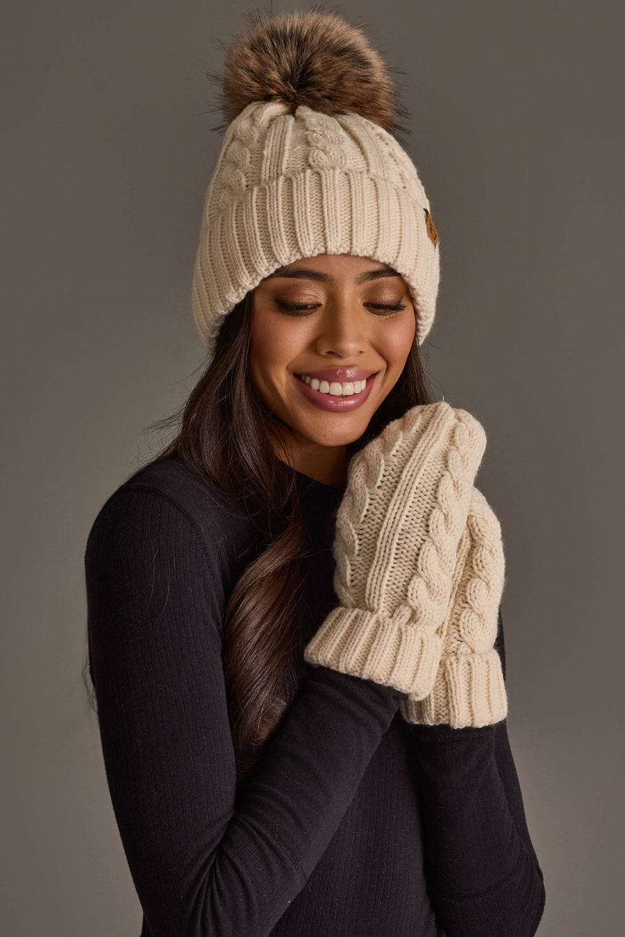 A smiling woman wearing the Panache Apparel Collection Cream Cable Knit Design Pom Hat, paired with matching knit mittens and a black top, stands against a plain background.