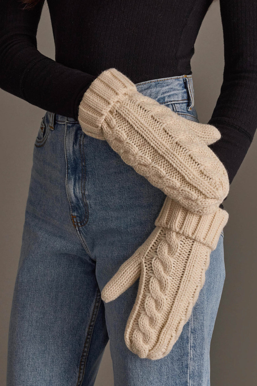 A person wearing a black ribbed long-sleeve shirt, blue jeans, and Panache Apparel Collection’s Cream Cable Knit Design Mittens stands against a plain backdrop.