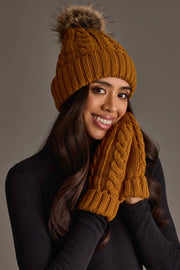 A woman smiles, holding her gloved hands to her cheek as she models the Panache Apparel Collection’s Mustard Cable Knit Design Pom Hat, featuring a cozy fur pom-pom, paired with matching knit gloves. Her long, wavy hair cascades over a black sweater.
