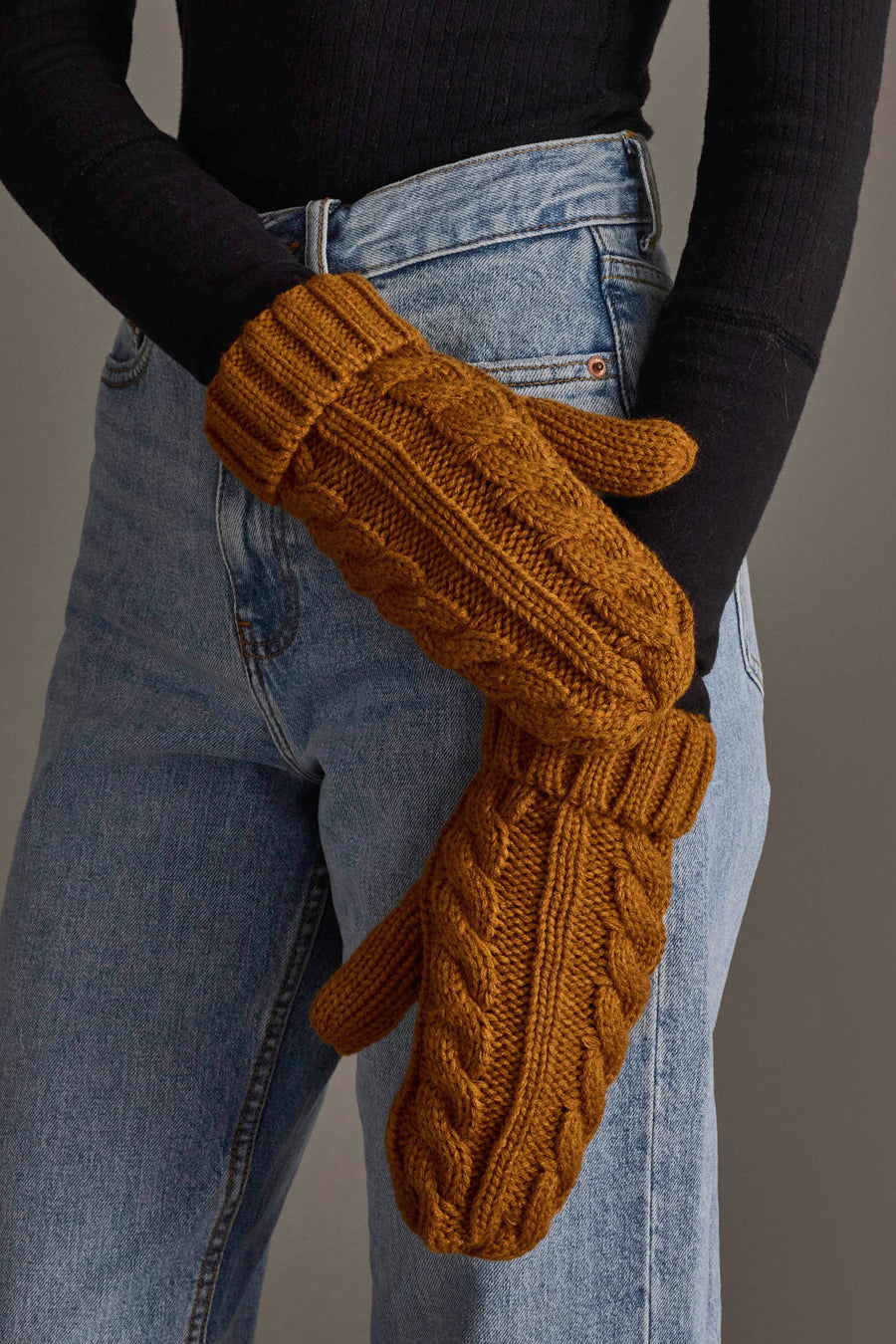 A person wearing a black long-sleeve shirt and blue jeans is shown with their hands crossed, wearing Panache Apparel Collection’s Mustard Cable Knit Design Mittens.