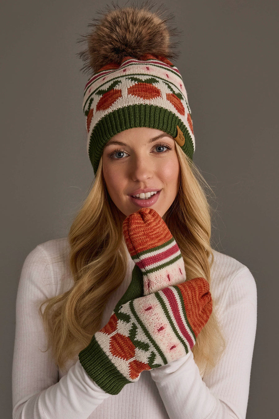 A woman in a white top wears the Olive, Cream, Rust & Pink Patterned Pom Hat and matching mittens from Panache Apparel Collection, smiling at the camera.