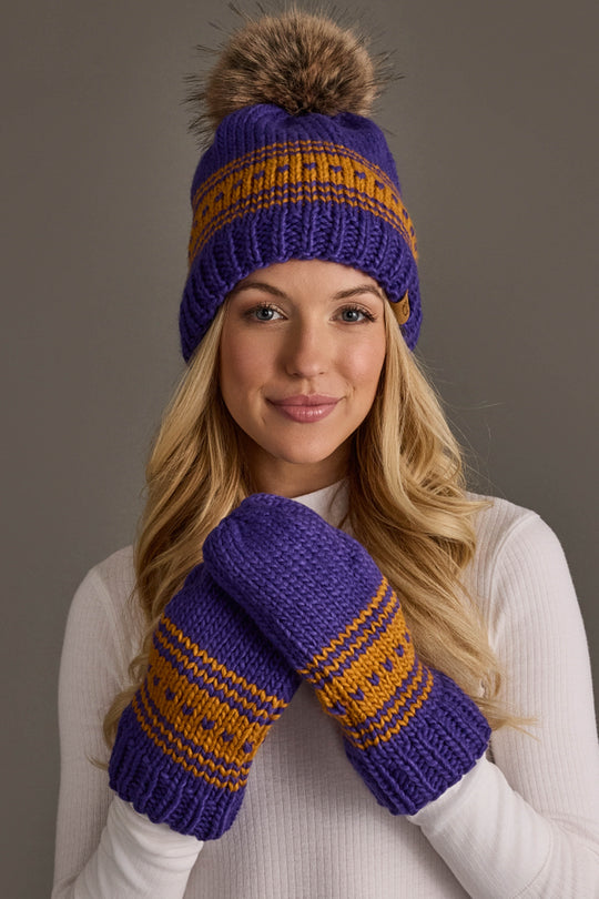 A person wearing the Panache Apparel Collection’s Purple & Gold Patterned Knit Pom Hat, paired with matching purple and orange knit mittens, smiles at the camera in a white turtleneck.