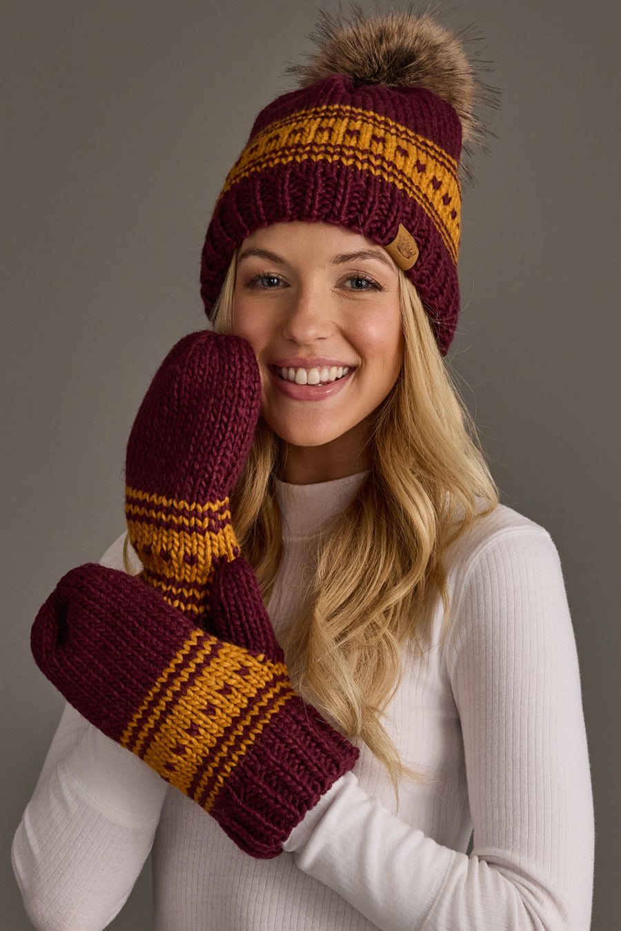 A woman in a white turtleneck sweater wears the Maroon & Gold Patterned Knit Pom Hat by Panache Apparel Collection, along with matching mittens, as she smiles at the camera against a neutral background.