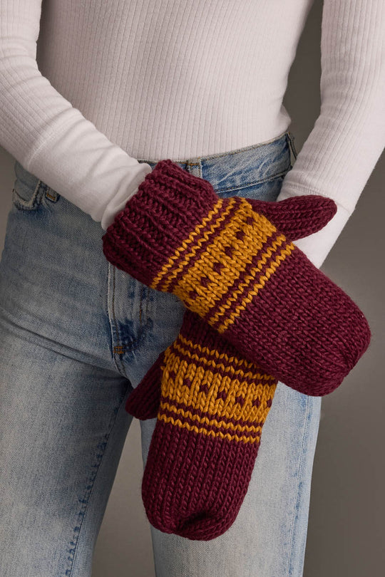 A person wearing a white ribbed top and blue jeans crosses their arms in front of their body while holding the Maroon & Gold Patterned Knit Mittens from Panache Apparel Collection.