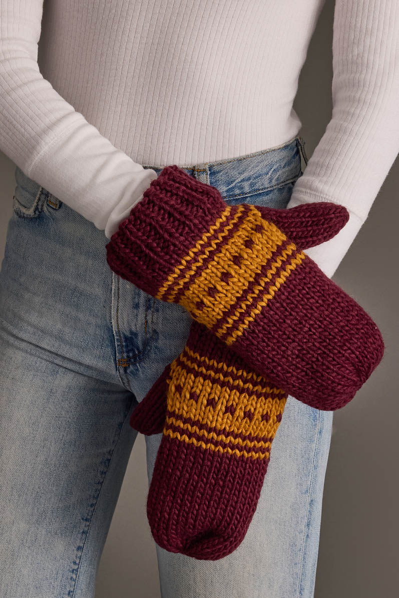 A person wearing a white ribbed top and blue jeans crosses their arms in front of their body while holding the Maroon & Gold Patterned Knit Mittens from Panache Apparel Collection.