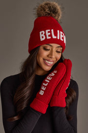 A person wearing a Red Believe Knit Pom Hat and matching mittens, both featuring the word BELIEVE in white lettering on a red knit fabric with a faux fur pom-pom.