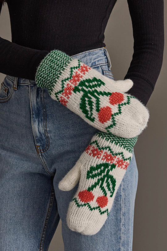 A person in a black top and blue jeans wears the Off White Cherry Pattern Mittens from Panache Apparel Collection, knit mittens that are fleece lined for extra warmth and adorned with green, red, and pink cherry and leaf designs.