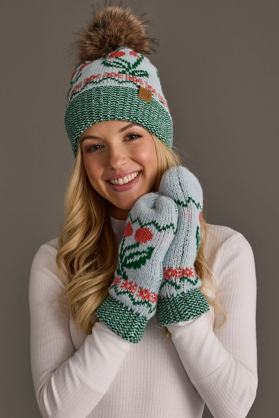 Smiling woman in a light blue knit hat with a cherry pattern and pompom from Panache Apparel Collection, wearing a white sweater.