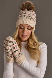 A woman wearing the Panache Apparel Collection Cream & Pink Floral Pattern Pom Hat, paired with matching mittens, looks down while dressed in a white turtleneck against a neutral background.