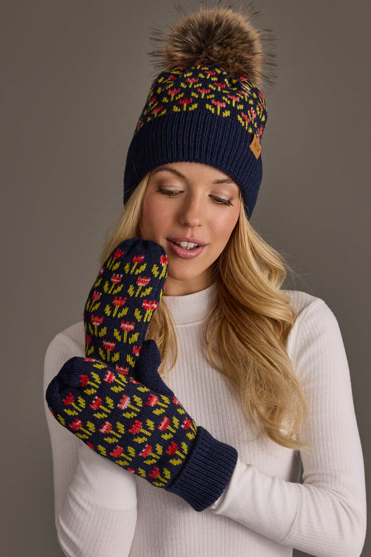 A woman wearing a white sweater and the Panache Apparel Collection Navy Small Floral Pattern Knit Pom Hat stands against a gray background, pairing it with matching patterned mittens featuring red, green, and yellow designs.