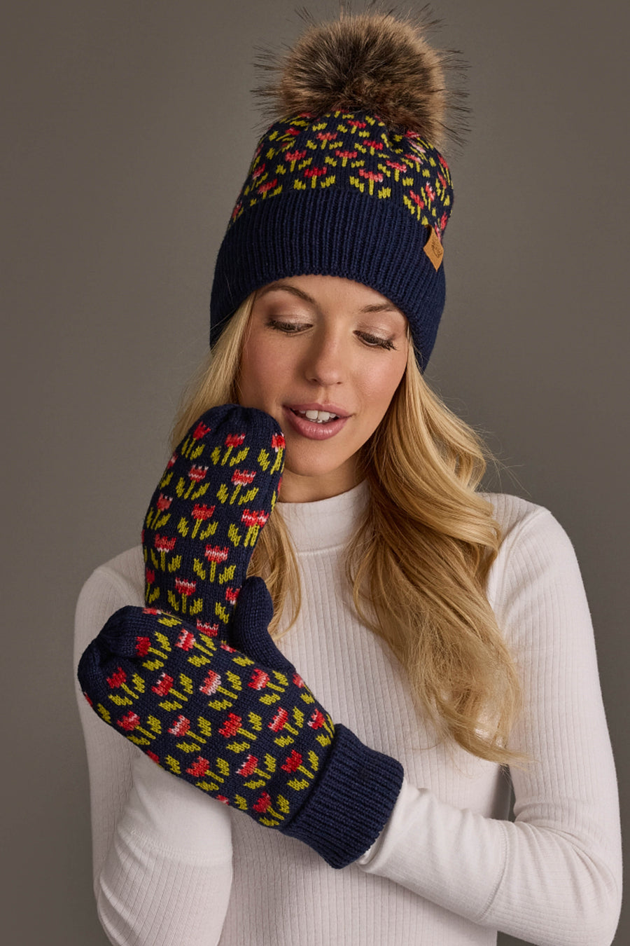 A woman wearing a white sweater and the Panache Apparel Collection Navy Small Floral Pattern Knit Pom Hat stands against a gray background, pairing it with matching patterned mittens featuring red, green, and yellow designs.