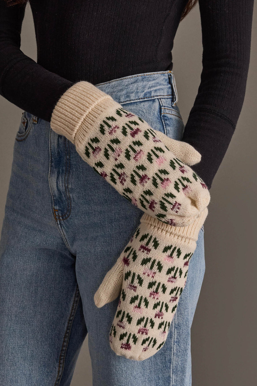 Wearing a black long-sleeve top and blue jeans, the person showcases the Cream Small Floral Pattern Knit Mittens from Panache Apparel Collection, displaying one mitten by holding it in their other hand.