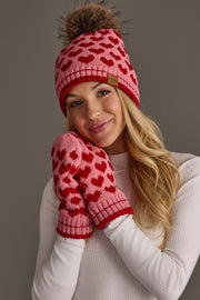 A woman smiles softly with her hands near her face, wearing a white sweater and the Panache Apparel Collection Pink & Red Heart Pattern Pom Hat, against a plain background.