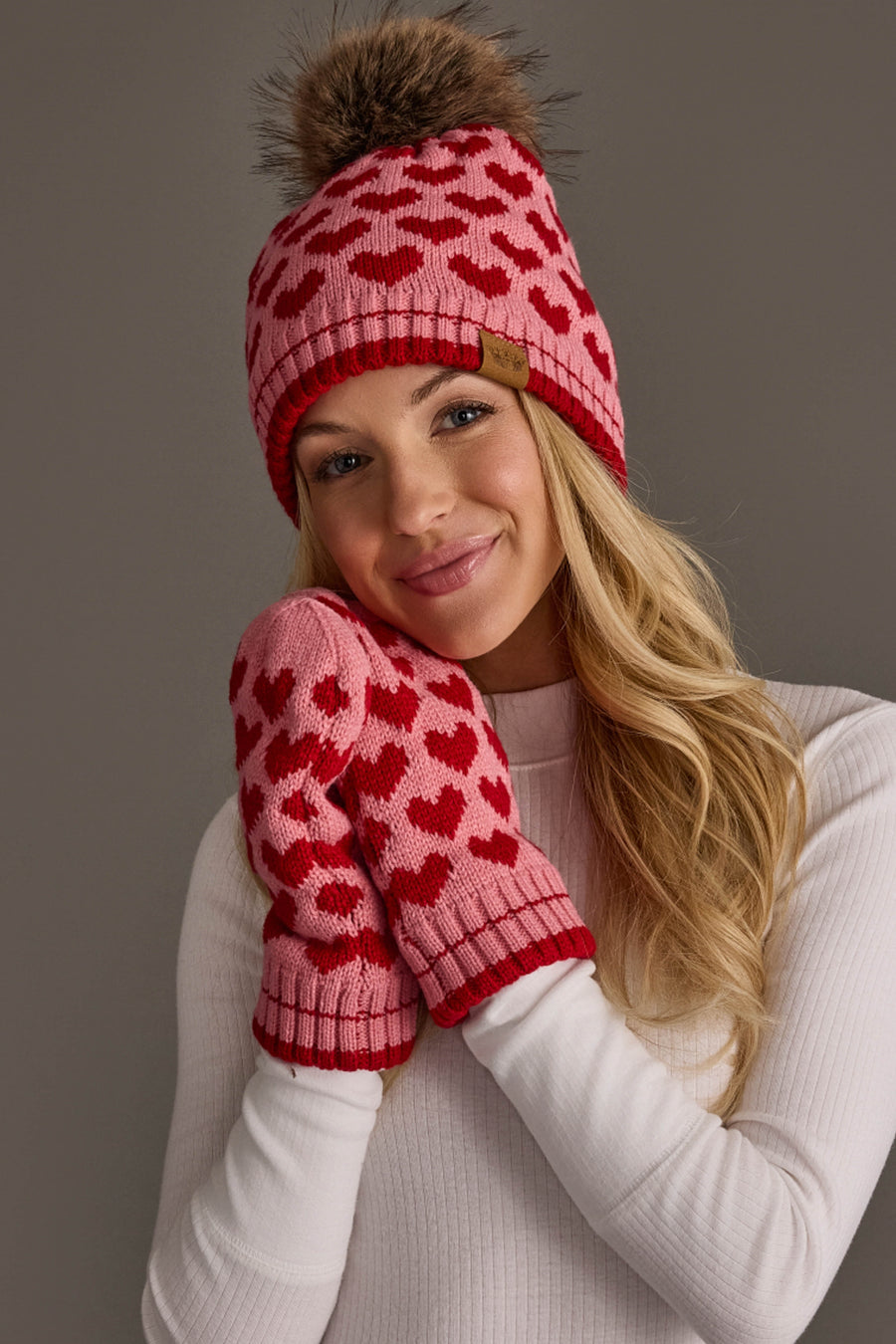 A woman smiles softly with her hands near her face, wearing a white sweater and the Panache Apparel Collection Pink & Red Heart Pattern Pom Hat, against a plain background.