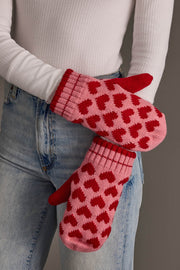 A person in blue jeans and a white ribbed top holds the Pink & Red Heart Pattern Mittens from Panache Apparel Collection.