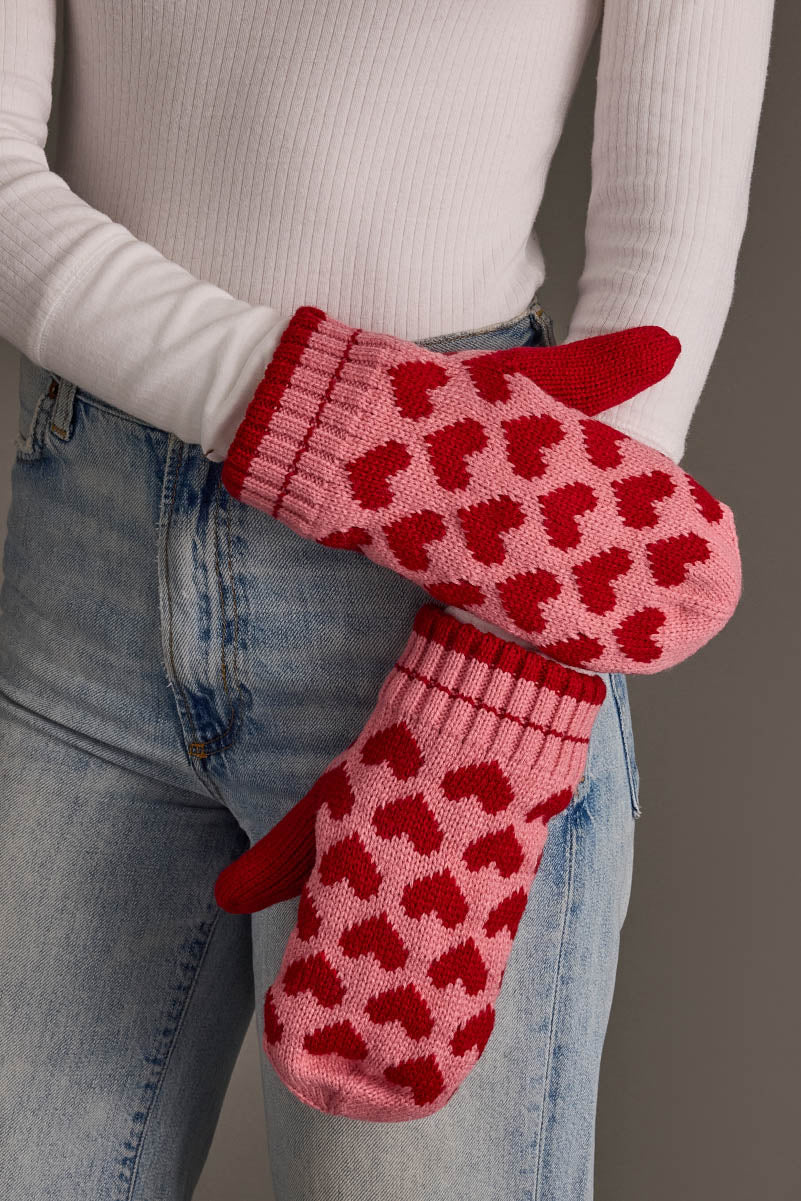 A person in blue jeans and a white ribbed top holds the Pink & Red Heart Pattern Mittens from Panache Apparel Collection.