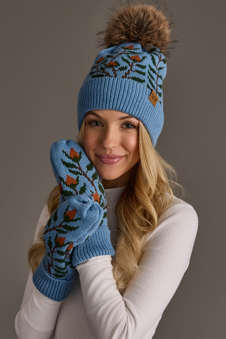 A woman in a white sweater wears the Panache Apparel Collection Blue Floral Pattern Knit Pom Hat, paired with matching mittens, and smiles at the camera against a neutral background.