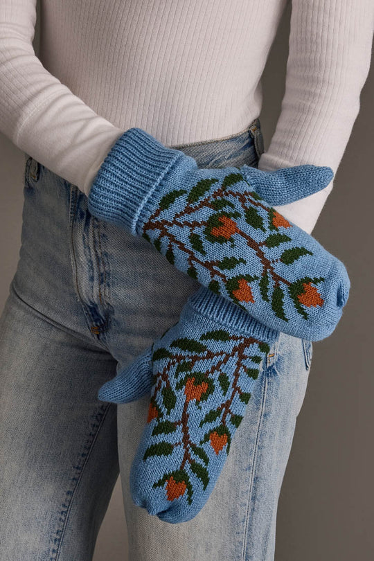 A person wearing Panache Apparel Collection’s Blue Floral Pattern Knit Mittens, featuring a green leaf and orange berry design, paired with a white ribbed sweater and light blue jeans, stands with their arms crossed.