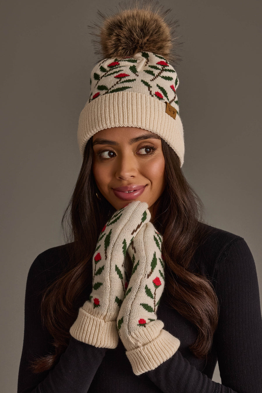 A woman smiles softly with her hands near her face, wearing the Panache Apparel Collection Cream Floral Pattern Knit Pom Hat.