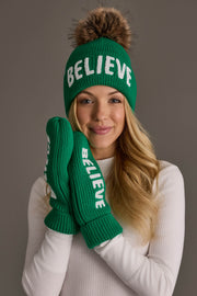 A woman wears a green acrylic knit beanie with a faux fur pom-pom and matching Green Believe Knit Mittens, both featuring the word BELIEVE in white lettering and fleece lining.