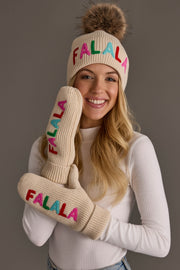 A woman wears the Beige Fa La La Pom Hat and matching mittens, both in beige knit with faux fur pompom and colorful FALALA embroidery.