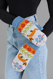Two people wearing light blue jeans and a black sweater, holding hands. They are wearing the Panache Apparel Collection Blue Fox Pattern Knit Mittens, featuring colorful animal, flower, and geometric designs in shades of orange, blue, red, and green.
