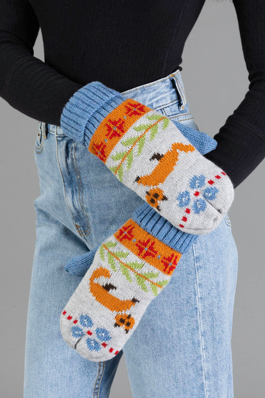 Two people wearing light blue jeans and a black sweater, holding hands. They are wearing the Panache Apparel Collection Blue Fox Pattern Knit Mittens, featuring colorful animal, flower, and geometric designs in shades of orange, blue, red, and green.