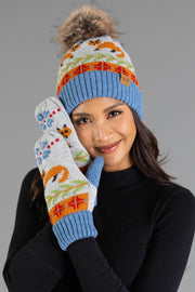 A woman in a black top smiles, wearing the Panache Apparel Collection’s Blue Fox Pattern Knit Pom Hat with a fur pom-pom, along with matching mittens featuring colorful fox and tree designs on a gray background.