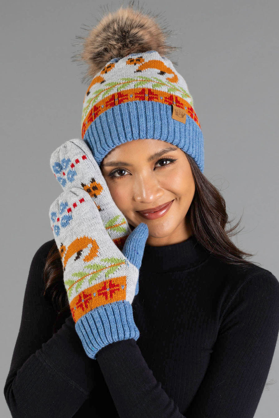A woman in a black top smiles, wearing the Panache Apparel Collection’s Blue Fox Pattern Knit Pom Hat with a fur pom-pom, along with matching mittens featuring colorful fox and tree designs on a gray background.