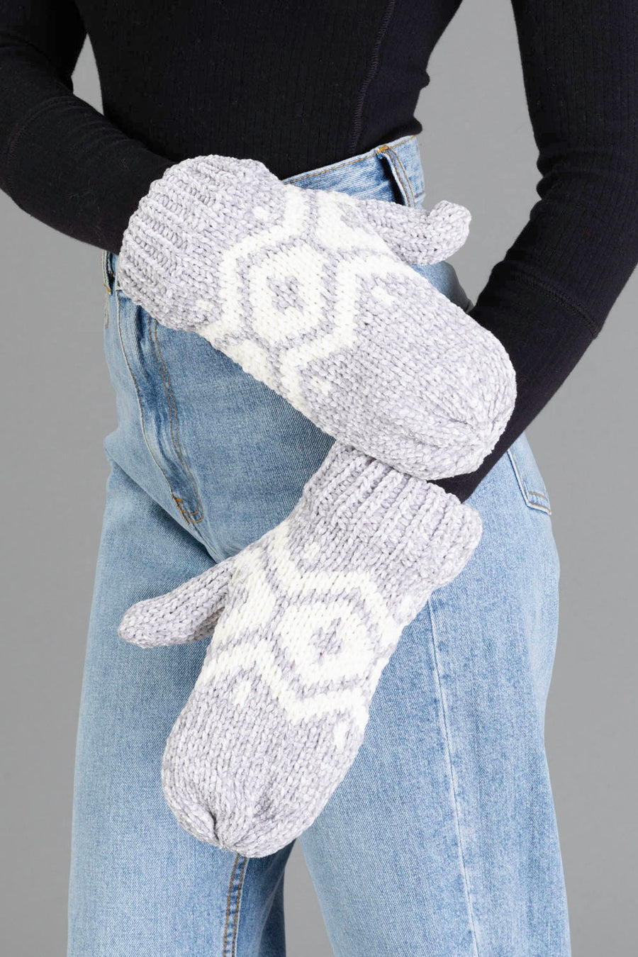 A person wears Panache Apparel Collection’s Gray & Cream Patterned Chenille Mittens with a black long-sleeve top and high-waisted blue jeans, arms crossed in front of their body against a plain gray background.