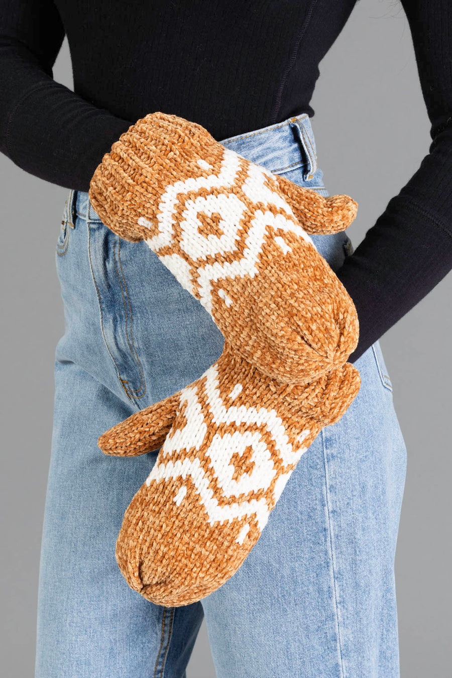 A person wearing blue jeans, a black long-sleeve top, and the Camel & Cream Patterned Chenille Mittens from Panache Apparel Collection stands against a gray background.