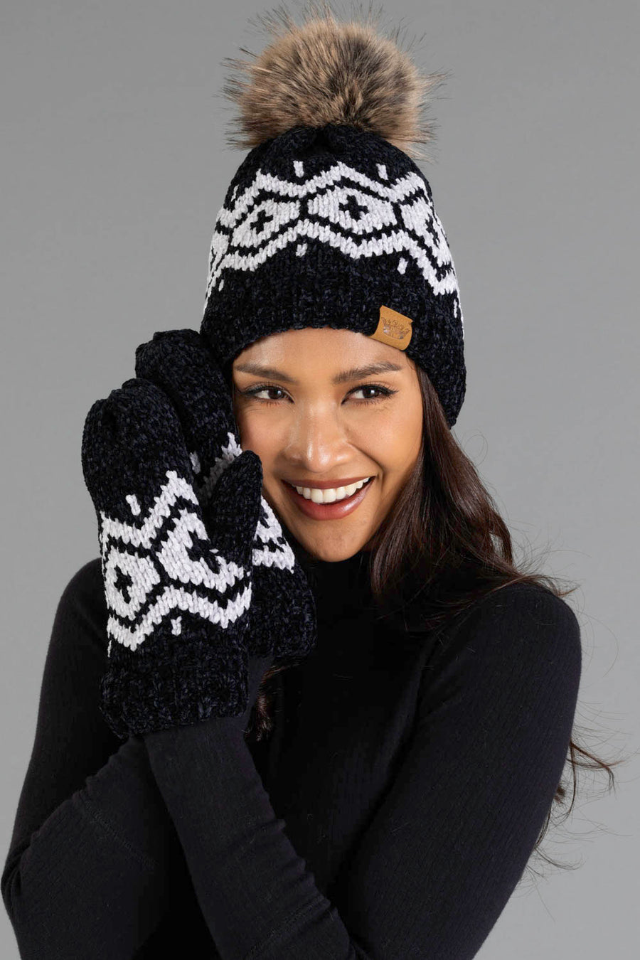 A woman smiles and holds her face with gloved hands, wearing the Panache Apparel Collection Black & Light Gray Patterned Chenille Pom Hat and matching mittens, paired with a black turtleneck against a gray background.
