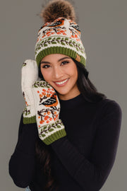 A smiling woman in a black top models the Olive & Cream Bird Pattern Knit Pom Hat by Panache Apparel Collection, featuring orange, black, and green patterns with a pom-pom on top, paired with matching mittens against a gray background.