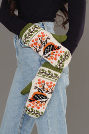 A person in blue jeans and a black top poses with hands in pockets, wearing Olive & Cream Bird Pattern Knit Mittens by Panache Apparel Collection, featuring bird and orange floral patterns with green cuffs and design accents.