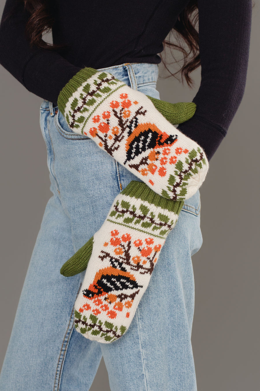 A person in blue jeans and a black top poses with hands in pockets, wearing Olive & Cream Bird Pattern Knit Mittens by Panache Apparel Collection, featuring bird and orange floral patterns with green cuffs and design accents.