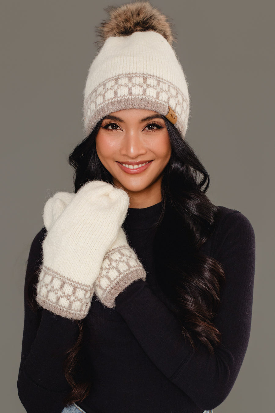 A woman with long dark hair, dressed in a black top, smiles at the camera while wearing the Ivory & Taupe Patterned Knit Pom Hat by Panache Apparel Collection and matching mittens, set against a neutral background.