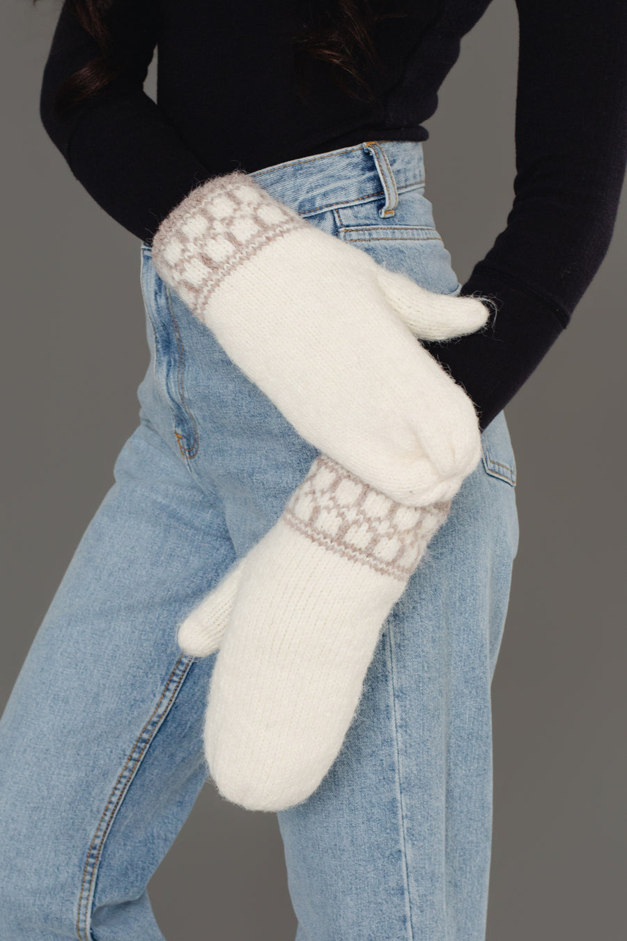 A person stands against a gray background wearing light blue jeans, a black long-sleeve top, and the Ivory & Taupe Patterned Knit Mittens from Panache Apparel Collection.