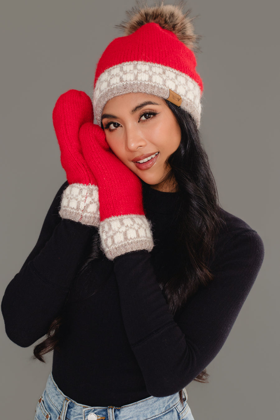 A woman smiles against a plain gray background, wearing the Panache Apparel Collection Red & Taupe Patterned Knit Pom Hat with a fur pom-pom and matching red mittens featuring gray and white trim. She pairs them with a black long-sleeve top and light blue jeans.