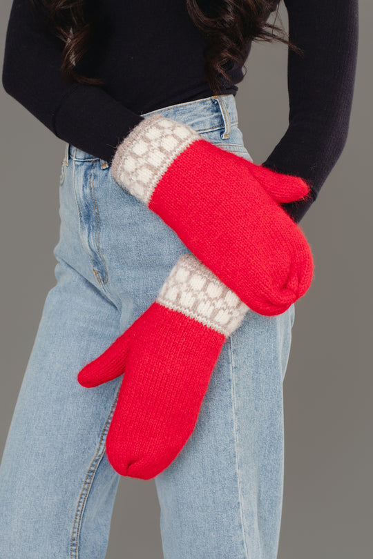 A person in light blue jeans and a black top crosses their arms against a gray background, wearing Panache Apparel Collection’s Red & Taupe Patterned Knit Mittens.