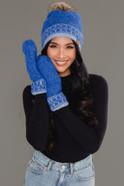 A woman smiles while wearing the Panache Apparel Collection’s Blue & Light Blue Patterned Knit Pom Hat and matching blue mittens, paired with a black long-sleeve top and light blue jeans, standing against a gray background.
